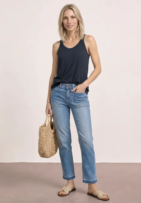 Vrouw in marineblauw hemdje, lichte spijkerbroek, sandalen en strooien tas voor neutrale achtergrond.