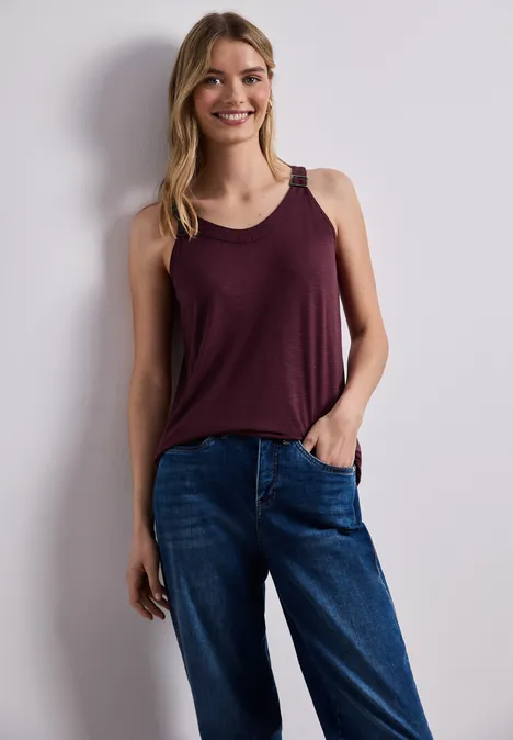 Frau steht entspannt vor einer weißen Wand, trägt bordeauxfarbenes Top und blaue Jeans.