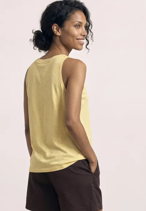 Vrouw van achteren in een geel mouwloos shirt en donkere short tegen lichte achtergrond.