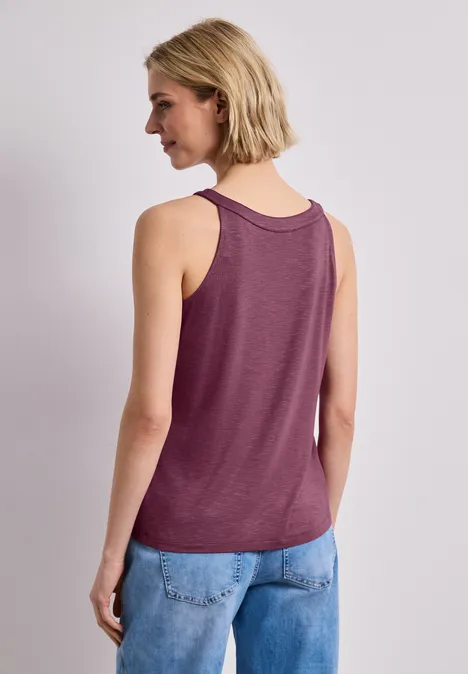 Rückansicht einer Frau im ärmellosen weinroten Top und blauen Jeans vor hellem Hintergrund.