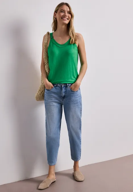 Frau steht vor neutralem Hintergrund in grünem Top, blauen Jeans, beigen Schuhen und Strohtasche.