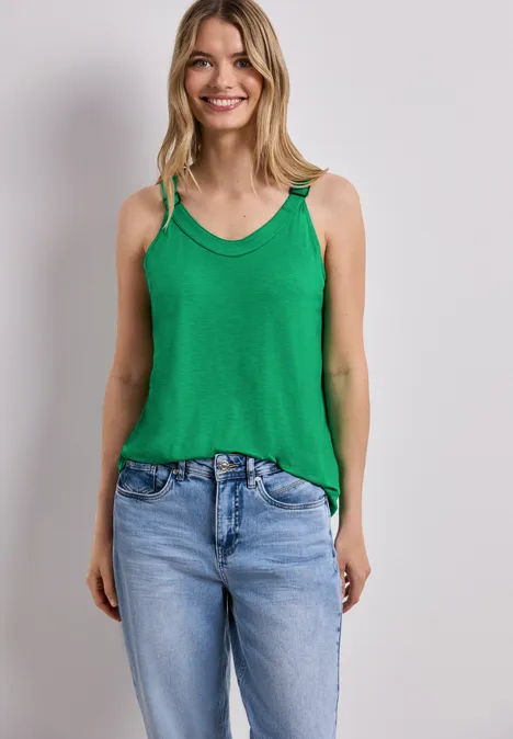 Frau trägt grünes Top und Jeans vor weißer Wand.