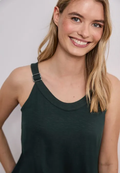 Frau mit langen blonden Haaren trägt ein dunkles ärmelloses Top vor neutralem Hintergrund.