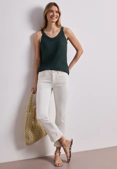 Frau mit grünem Top, weißen Jeans, Sandalen und Korbtasche lehnt an weißer Wand.