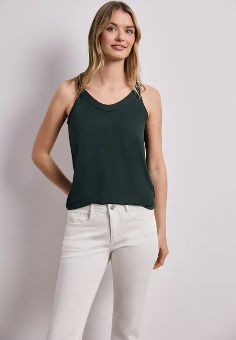 Frau in dunkelgrünem Top und weißen Jeans vor neutralem Hintergrund.