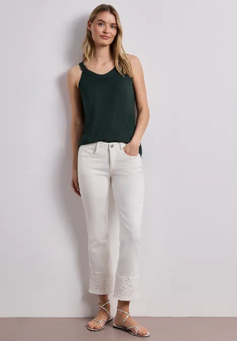 Junge Frau in schwarzem Top, weißen Hosen mit Spitzenbesatz und Sandalen vor hellem Hintergrund.