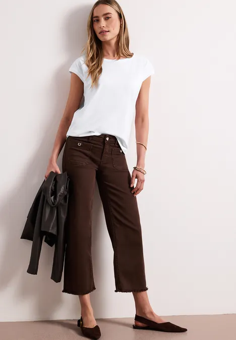 Vrouw in wit T-shirt en bruine culotte, met zwarte jas in hand, tegen een lichte muur.