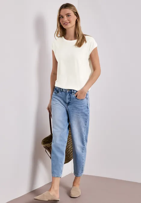 Frau trägt weißes T-Shirt, lockere blaue Jeans und Basttasche, steht lässig vor hellem Hintergrund.