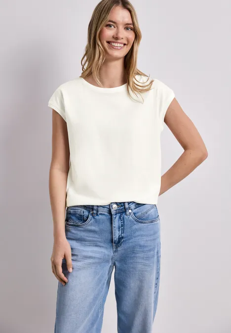 Eine Frau trägt ein weißes T-Shirt mit kurzen Ärmeln und blaue Jeans vor neutralem Hintergrund.