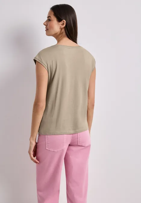 Vrouw van achteren in een beige top en roze broek tegen een witte achtergrond.