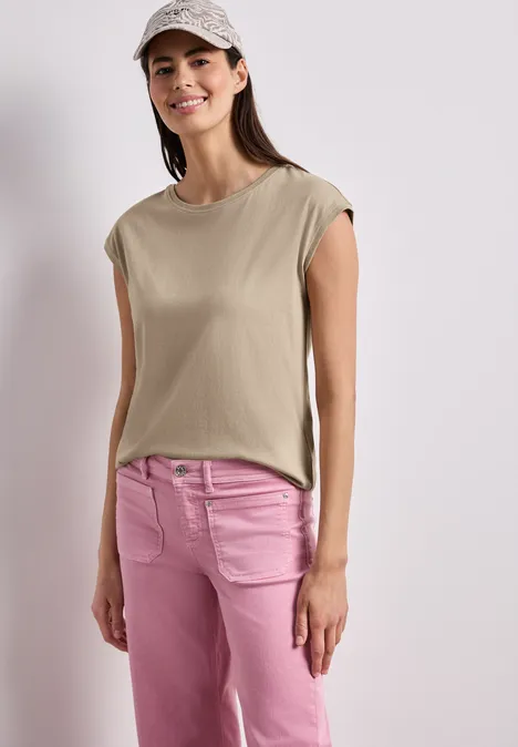 Een vrouw draagt een mouwloos beige topje en roze spijkerbroek tegen een neutrale achtergrond.