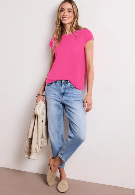Vrouw in roze T-shirt, losse blauwe jeans en beige loafers, leunend tegen een muur met een lichte jas in de hand.