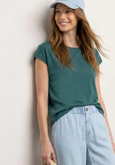 Eine Frau trägt ein grünes T-Shirt, helle Jeans und eine Kappe vor weißem Hintergrund.