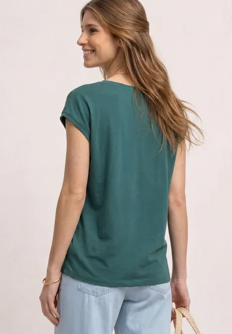 Frau von hinten in grünem T-Shirt und Jeans vor schlichtem, hellem Hintergrund.