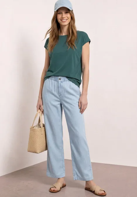 Frau mit grünem T-Shirt, hellen weiten Jeans, Sandalen und Strohtasche vor hellem, neutralem Hintergrund.