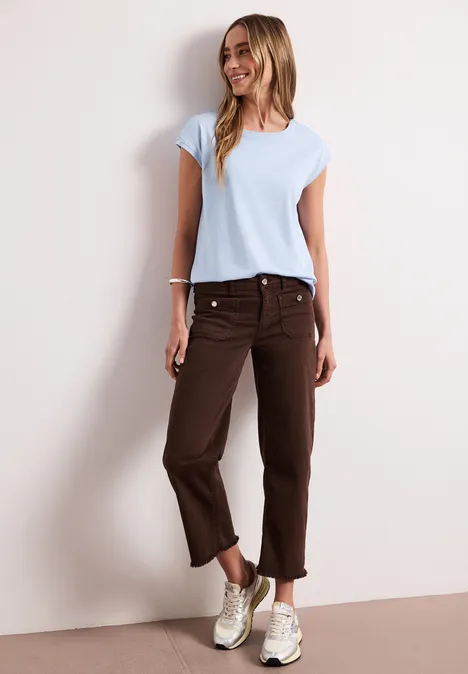 Frau in hellem T-Shirt, brauner 3/4-Hose und Sneakers vor neutralem Hintergrund.