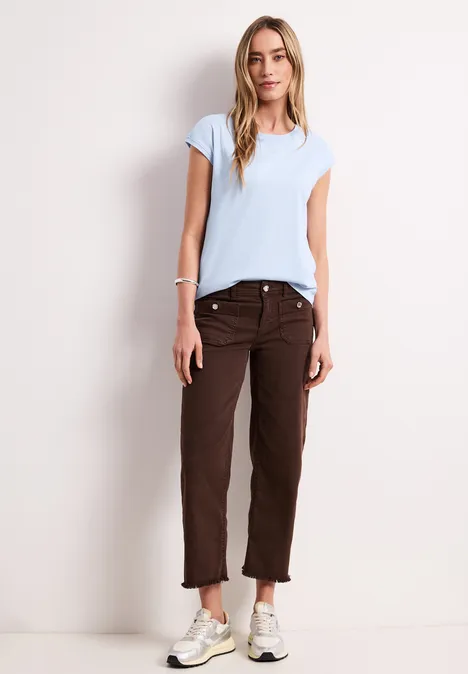 Frau mit hellblauem T-Shirt, braunen Hosen und weißen Sneakers vor weißer Wand.