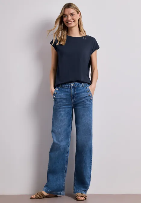 Frau trägt navyblaues T-Shirt und weite Jeans vor weißer Wand.