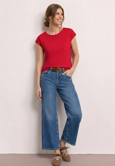 Shirt mit Boatneck und Deko-Detail salsa red Frau steht vor hellem Hintergrund, trägt rotes T-Shirt, weite Jeans, braunen Gürtel und Schuhe.
