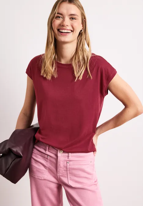 Vrouw in bordeaux t-shirt en roze jeans, houdt een tas vast voor een lichte achtergrond.