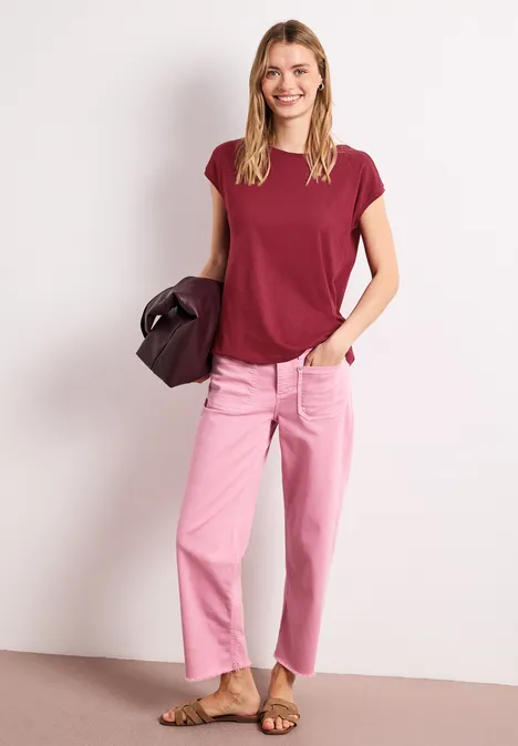 Vrouw in bordeauxrode top en roze wijde jeans, met tas in hand, poseert voor een neutrale achtergrond.