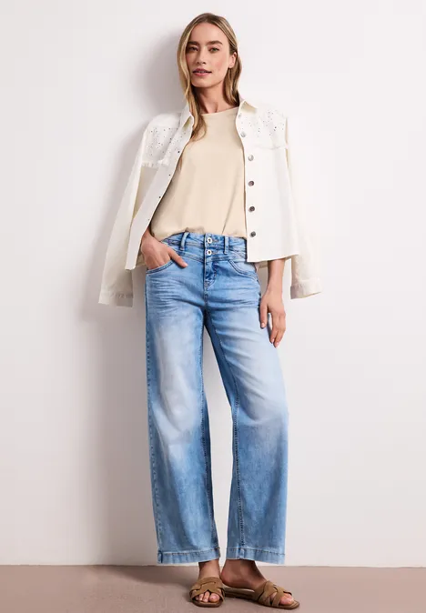 Een vrouw draagt een lichte jas nonchalant over de schouders, een beige top, wijdvallende jeans en sandalen.