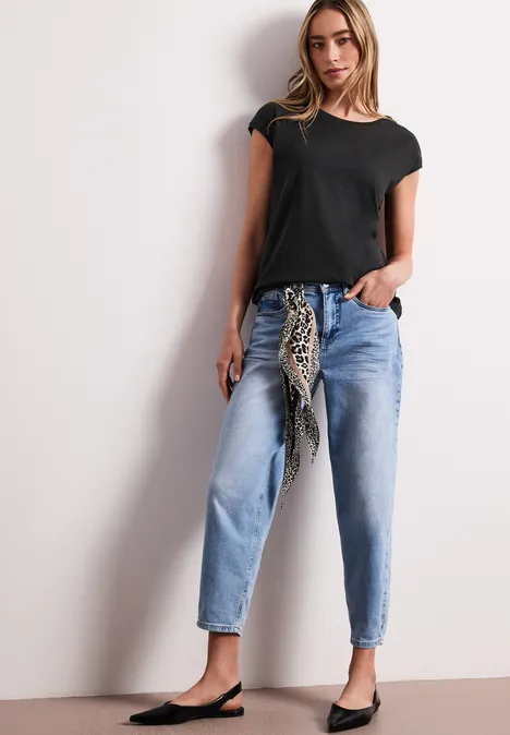 Vrouw in zwart T-shirt, lichtblauwe jeans, met een knoop-sjaal en platte schoenen.