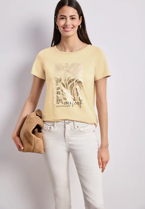 Frau trägt hellgelbes T-Shirt mit Pflanzenmotiv und weiße Hose, hält eine Handtasche vor neutralem Hintergrund.