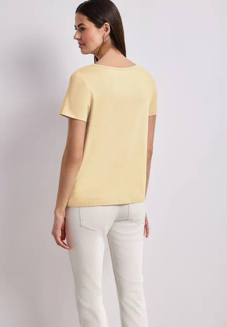 Frau von hinten in gelbem T-Shirt und weißer Hose vor hellem Hintergrund.