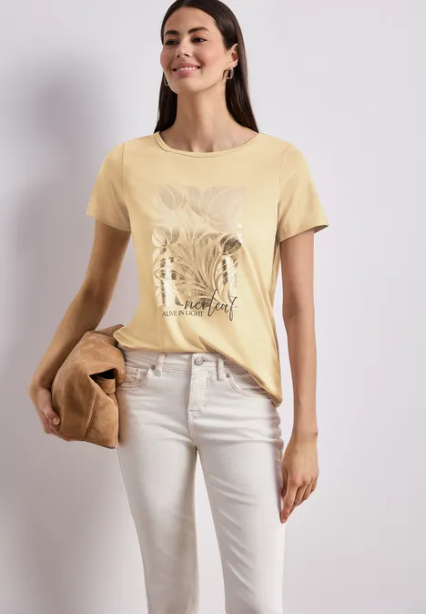 Frau in gelbem T-Shirt mit Pflanzenmotiv, hellen Hosen und brauner Tasche vor hellem Hintergrund.