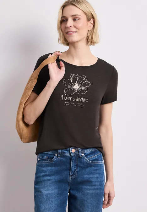 Frau trägt schwarzes T-Shirt mit weißer Lotusblüte und Schriftzug, kombiniert mit Jeans und Schultertasche.
