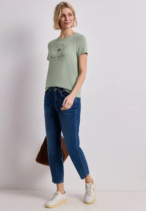 Frau in grünem T-Shirt, blauen Jeans, weißen Sneakern, steht lässig, trägt braune Tasche.