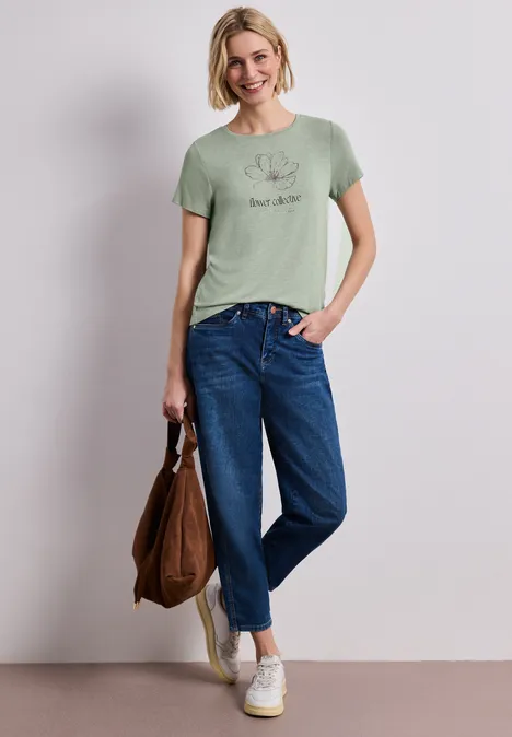 Frau trägt mintgrünes T-Shirt, blaue Jeans, weiße Sneakers und hält eine braune Tasche.