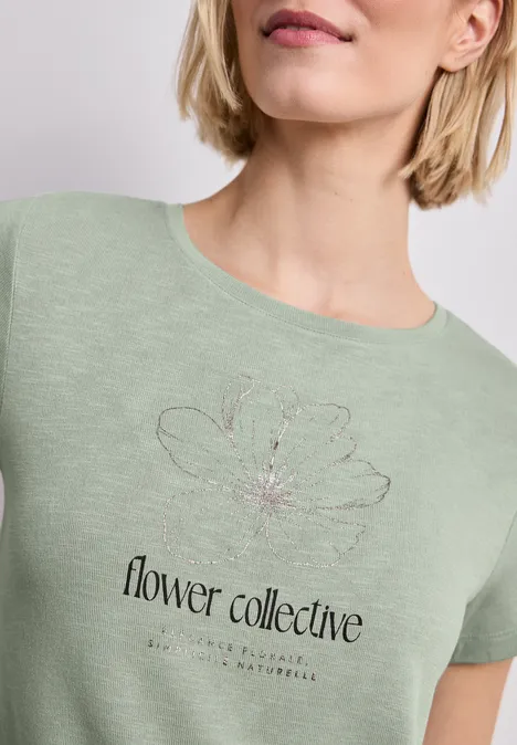 Pastellgrünes T-Shirt mit einfachem Blumendesign und Schriftzug 'flower collective' auf Brusthöhe.