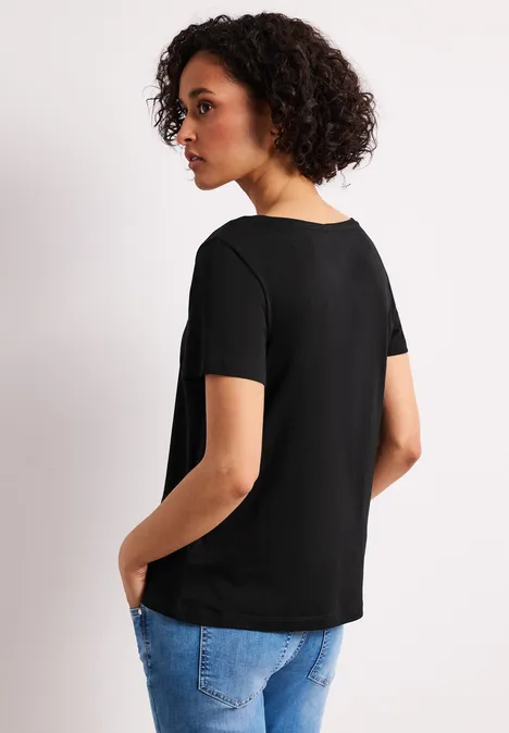 Eine Frau im Profil trägt ein schwarzes T-Shirt und blaue Jeans, Hände in den Taschen, lockige Haare.