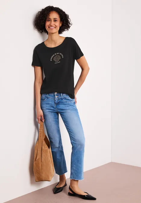 Frau mit lockigem Haar, in schwarzem T-Shirt, blauen Jeans, schwarzen Loafers und brauner Tasche.