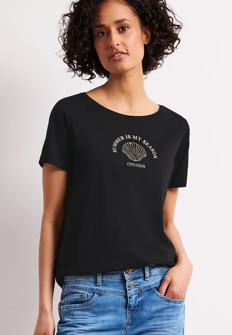 Frau trägt schwarzes T-Shirt mit Slogan und blaue Jeans vor hellem Hintergrund.