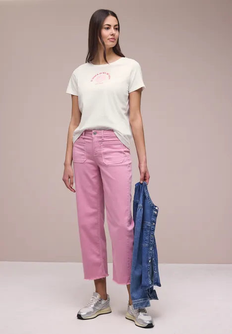 Frau trägt weißes T-Shirt, rosa Jeans und Sneaker, hält blaue Jeansjacke in der Hand.