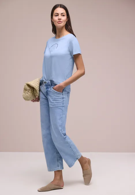 Eine Frau trägt hellblaues T-Shirt, lockere Jeans, Espadrilles und hält eine Strohtasche.