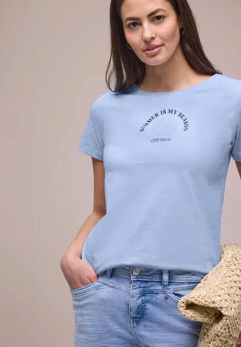 Frau in hellblauem Shirt und Jeans hält Strohtasche und posiert.