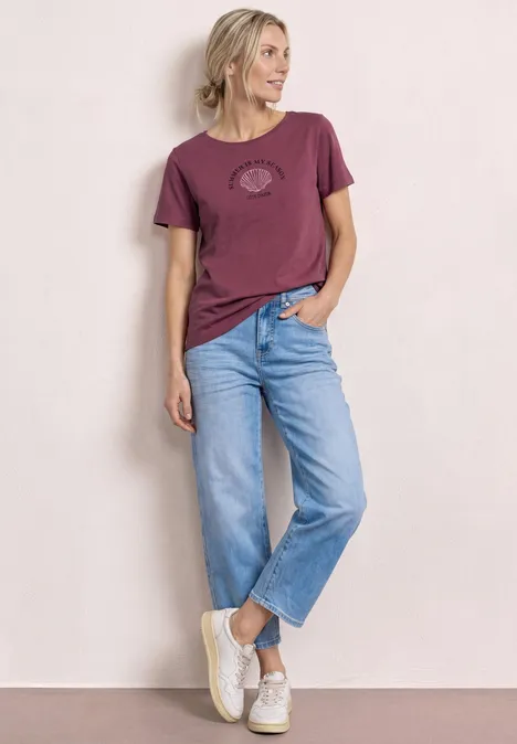 Vrouw met lichtgekleurde achtergrond in paars T-shirt, lichtblauwe jeans en witte sneakers.