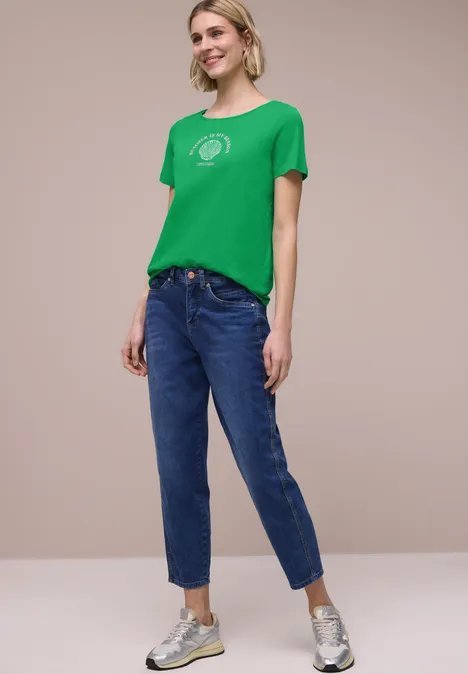 Vrouw met groen T-shirt, blauwe spijkerbroek en zilverkleurige sneakers voor een neutrale achtergrond