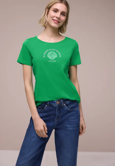 Vrouw in groen T-shirt met wit logo en blauwe jeans, staand voor een licht neutrale achtergrond.