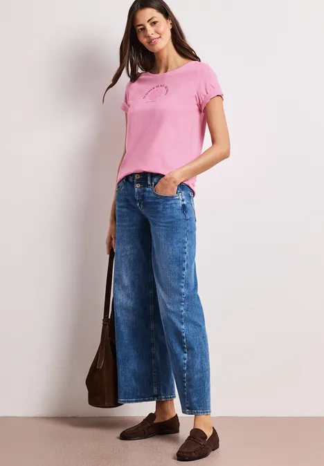 Frau in rosa T-Shirt, weiten Cropped-Jeans und braunen Loafers trägt eine braune Schultertasche.