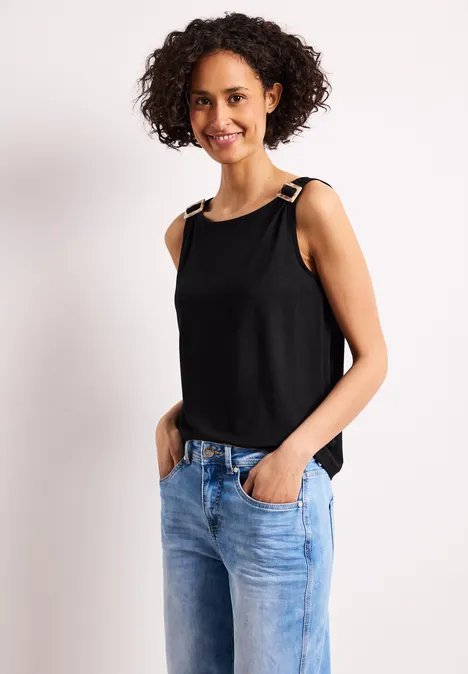 Eine Frau in schwarzem Trägertop und blauen Jeans, steht vor hellem Hintergrund.