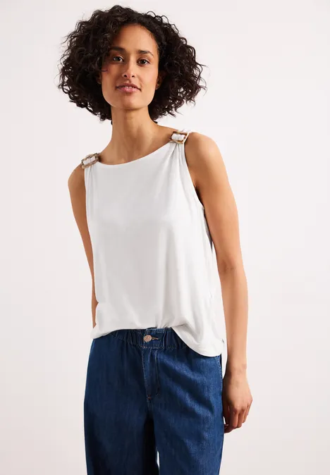 Frau mit lockigem Haar trägt ärmelloses weißes Top und blaue Jeans vor hellem Hintergrund.