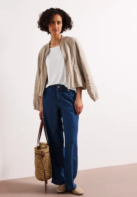 Frau trägt weißes T-Shirt, beige Jacke, weite Jeans, Korbtasche und beigen Slipper.