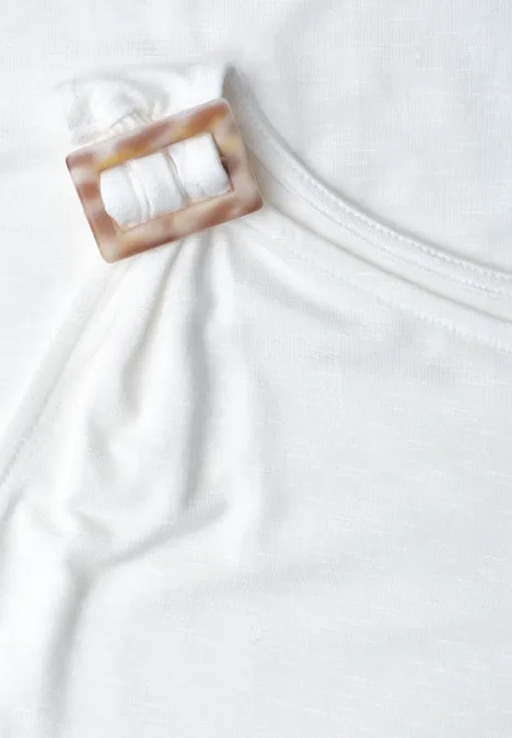 Nahaufnahme eines weißen Tops mit einem eckigen, hornfarbenen Schnallen-Detail am Träger.