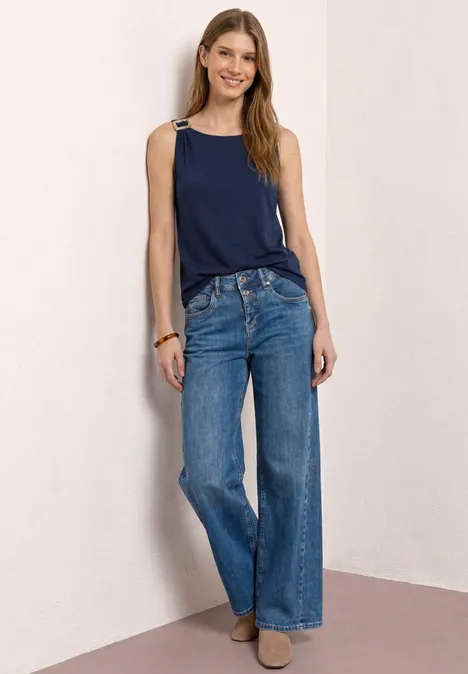 Frau lehnt an Wand, trägt dunkelblaues Top, weite Jeans und hellbraune Stiefeletten vor schlichtem Hintergrund.