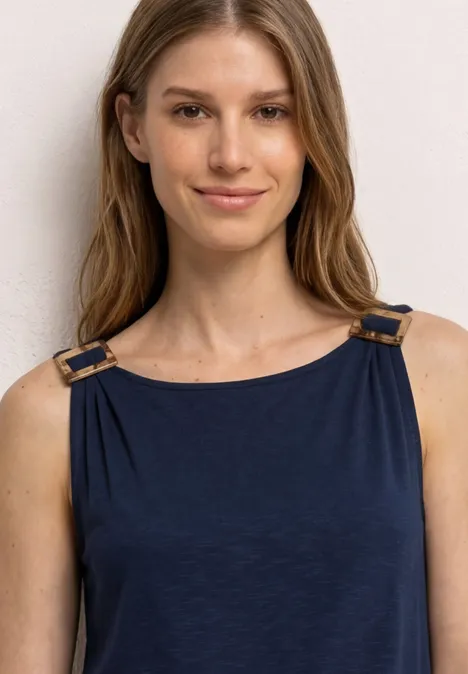 Eine Frau mit unscharfem Gesicht trägt ein ärmelloses blaues Top mit goldenen Schnallen.